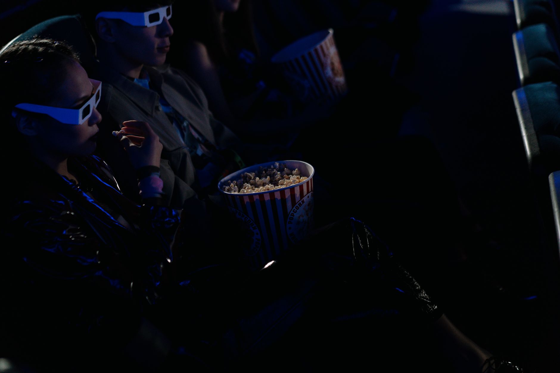 Audience members wearing 3D glasses and holding a popcorn bucket seated in a dark cinema auditorium.
