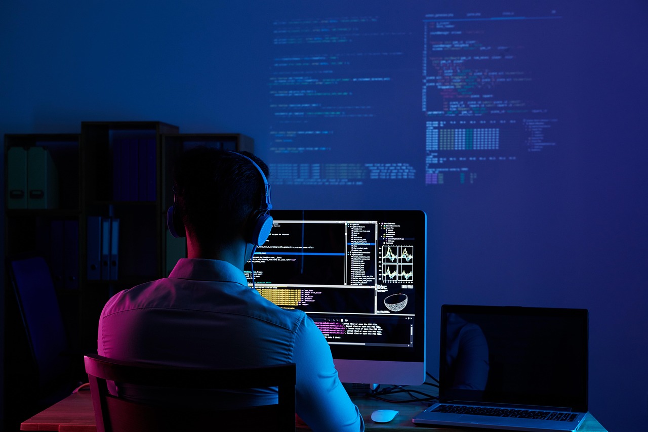 Developer seen from behind at a desk, facing several screens full of code in a dark room