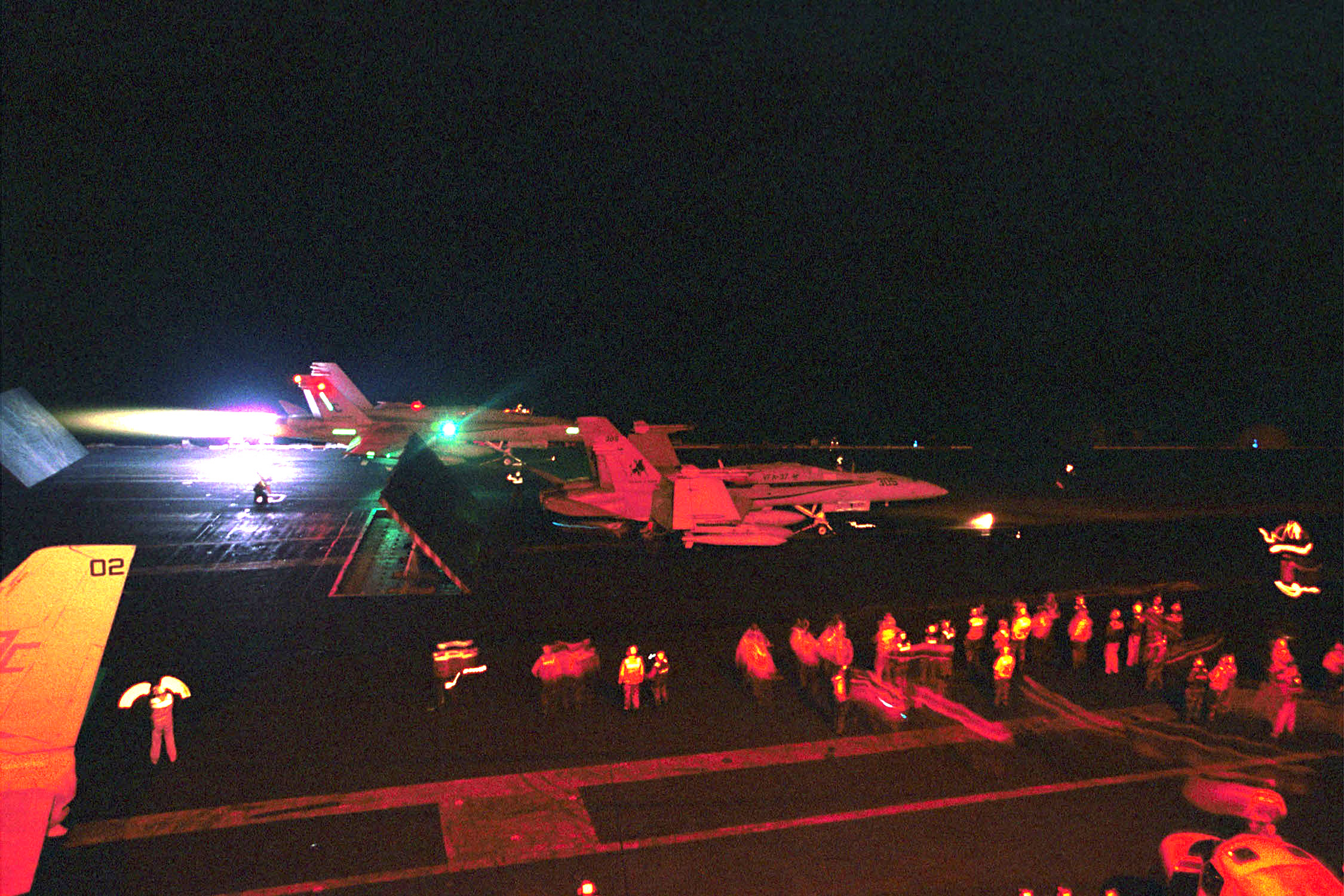 Fighter jets and deck crew on an aircraft carrier flight deck at night, lit in red and orange tones.