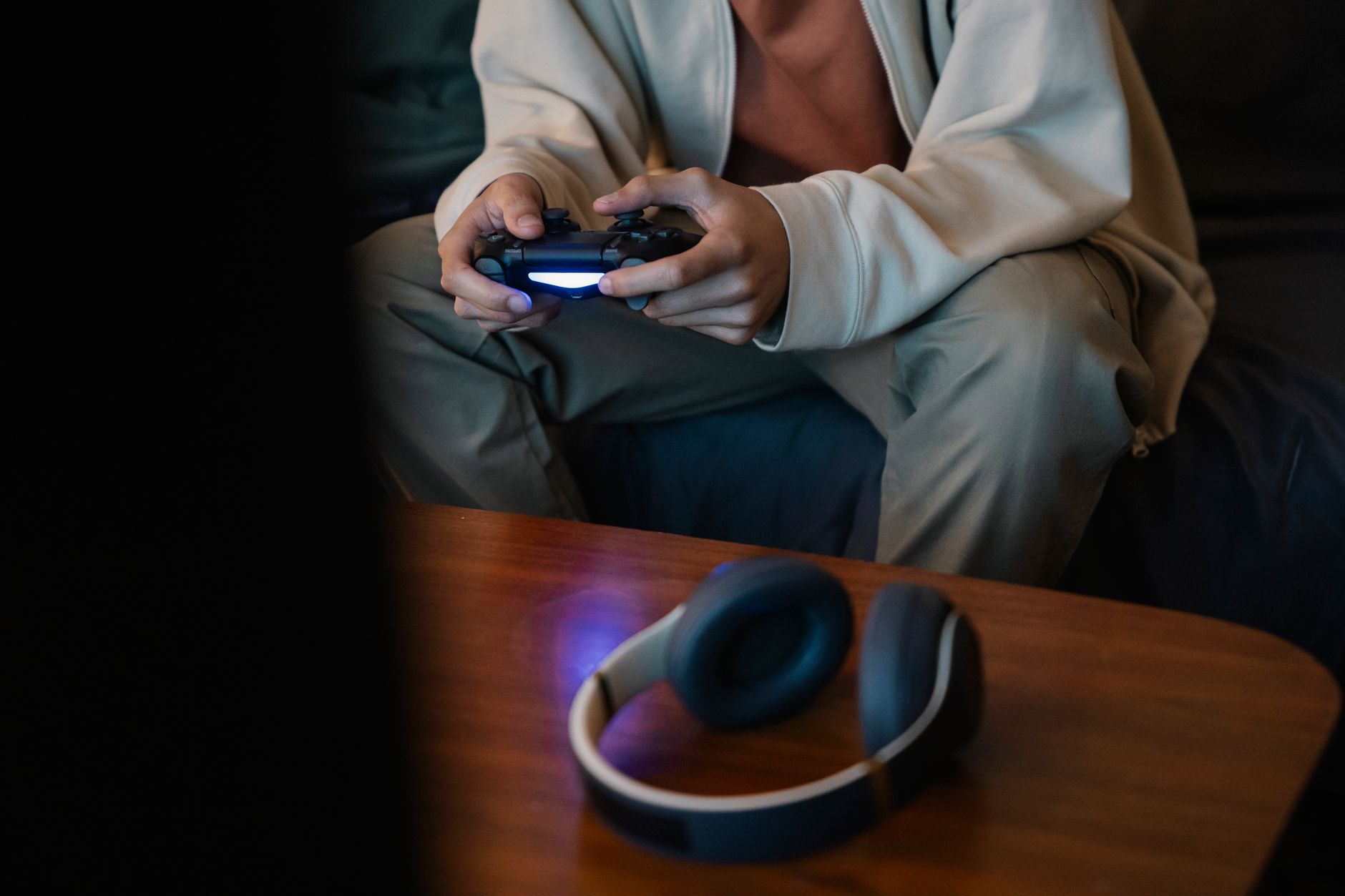 Person seated on a sofa in a dim room holding a gamepad with a blue light, with headphones on a wooden table in front.