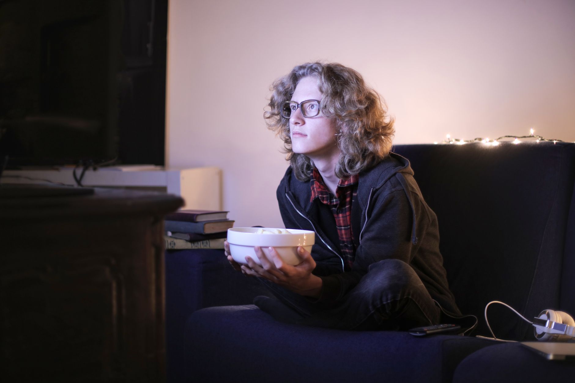 A person with glasses sits on a sofa holding a bowl while looking attentively at a television screen.