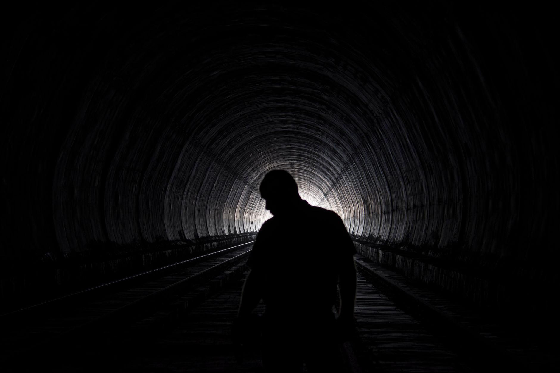 Silhouette of a person with head bowed standing between rails in a dark curved railway tunnel, with light at the far end.