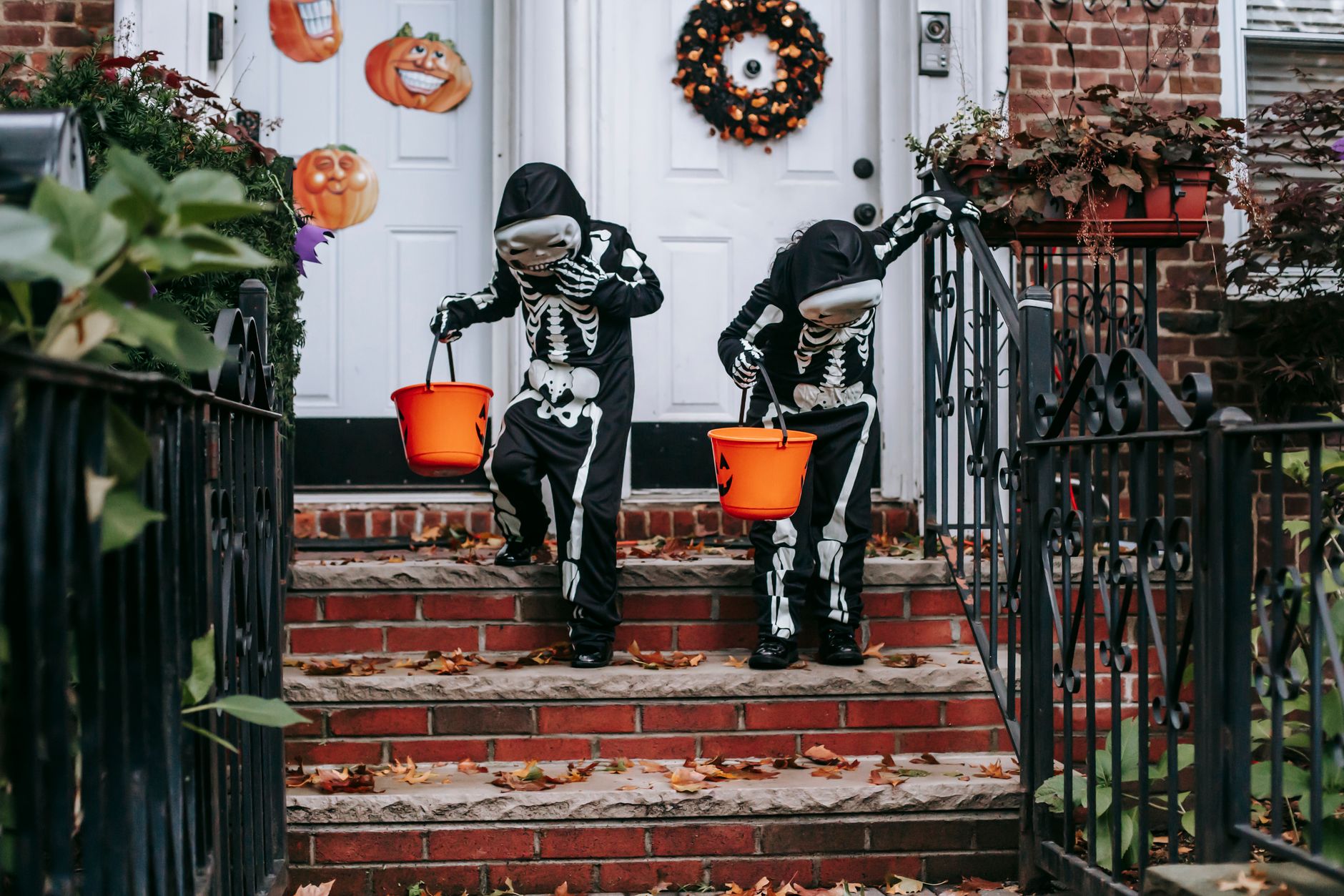 Two children in skeleton costumes carry orange pumpkin buckets down brick front steps decorated for Halloween.